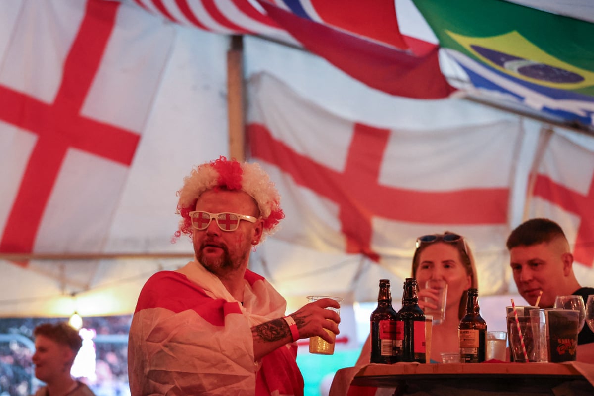 A fan wearing a wig and glasses in England colours and draped in the St George’s flag holds a pint alongside a table of drinks with two other fans