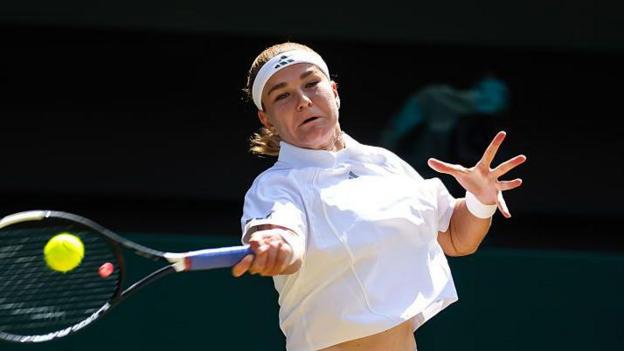 Karolina Muchova, wearing white tennis top and a white headband, hits a forehand as her face shows the strain of playing the shot