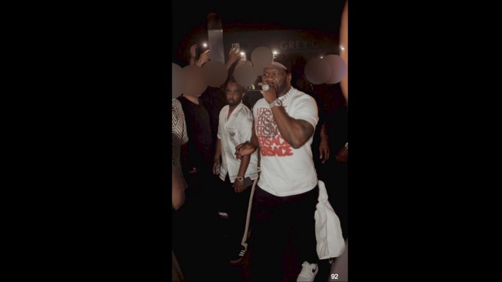 Hassan, wearing a silky button up shirt, stands next to 50 Cent, who is holding a microphone.