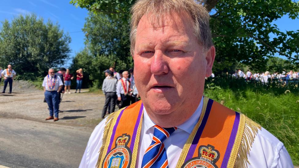 David Mahon, a man with short, fair hair, looks to the camera during a BBC interview on a very sunny day in rural Donegal. He is wearing a white shirt, a red. white and blue striped tie and an orange sash with a gold fringe.