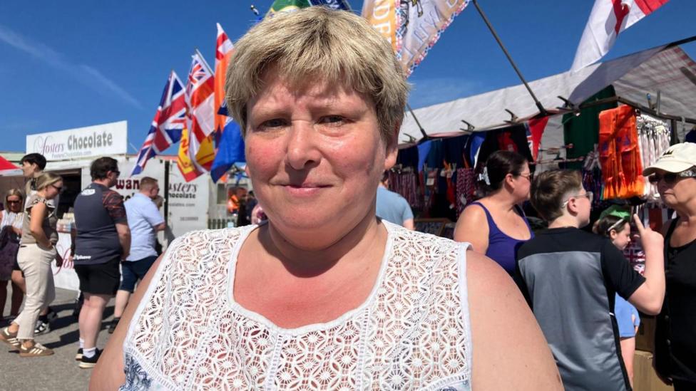 A woman with short light hair wearing a white and blue top looks at the camera on a sunny day. She is standing in front of a food stall. There are crowds of people in the background and row of flags which including the Union flag.