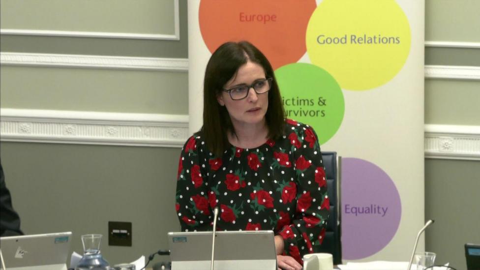 Alliance Party MLA Paula Bradshaw at the Executive Office committee. She has shoulder-length dark hair and is wearing glasses. She is wearing a dark dress with a red rose pattern on it and sitting behind a desk with tablet computer folded in front of her. Behind her is a pull-up banner with words such as