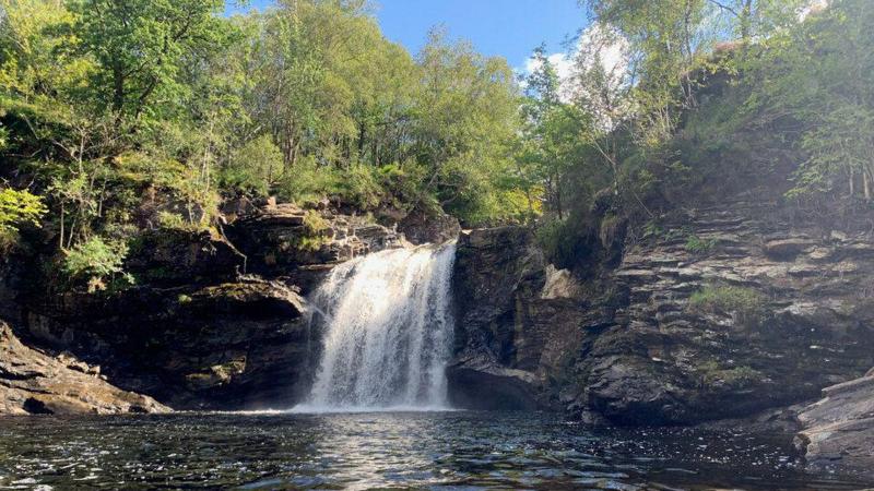 A small waterfall cascades over a rocky cliff into a clear, calm pool surrounded by steep rock walls and dense green trees. Sunlight filters through the foliage, reflecting on the water's surface and illuminating the layered stone formations along the shoreline.
