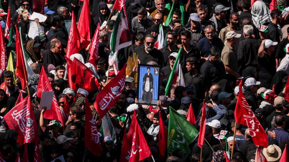 Mourners gather for the burial of Iranian Supreme Leader Ali Khamenei in Mashhad. Many are carrying red flags with Arabic script and in the centre a man holds a picture of a sitting Khamenei, with his son Mojtaba standing above him.