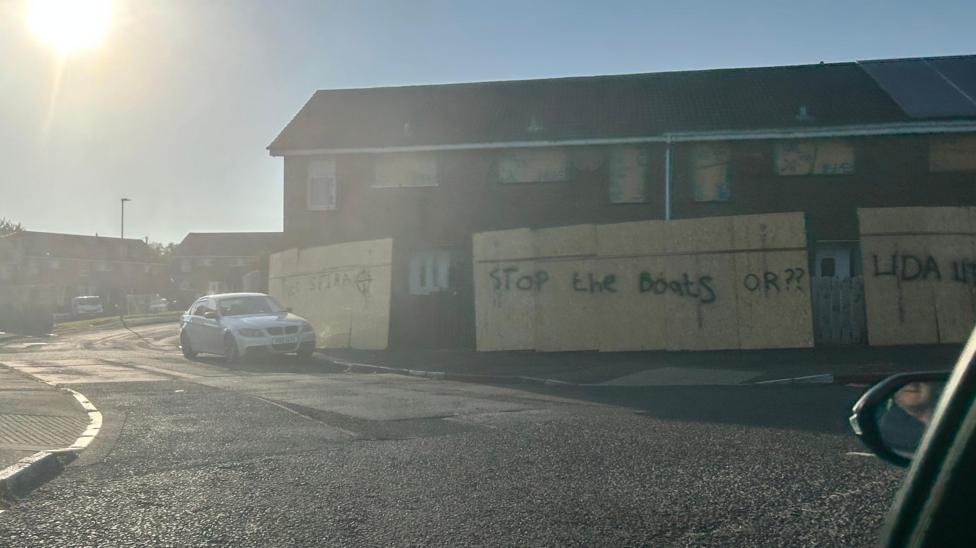 Hoarding outside a set of terrace houses sprayed with graffiti reading: STOP THE BOATS'. A car is parked to the left.