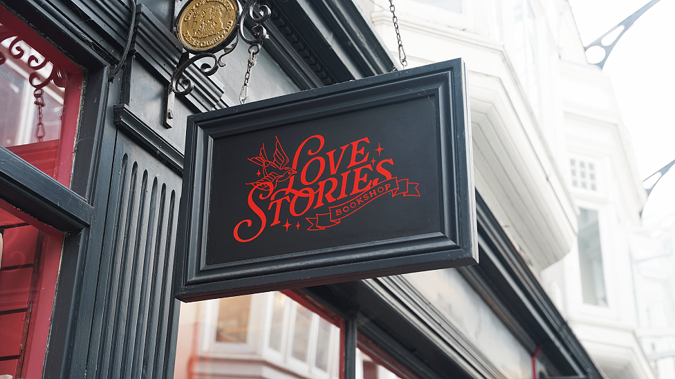 A photo of the Love Stories sign painted in dark grey-green, with red painted lettering. The sign is rectangular and hangs from a wrought iron bracket. The top of the windows can be seen, while above the upper floor windows and the covered skylight of the arcade with metal strut supports can be seen.