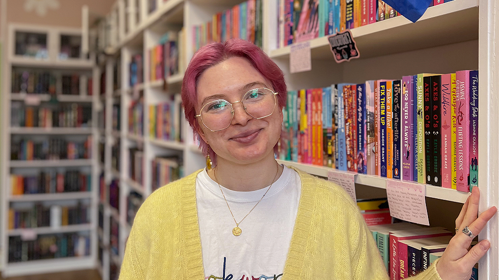 Caden Armstrong is a white women in her 20s. She has pink dyed short to neck-length hair, wears large wire-framed glasses with long dangly gold earrings ending in discs and a small gold medallion necklace. She wears a white t-shire with the top of some writing visible and a yellow cardigan over it. Her left hand rests against a bookshelf and has two silver rings visible. Behind her are rows of white bookshelves with books filling them, slightly out of focus as the background recedes.