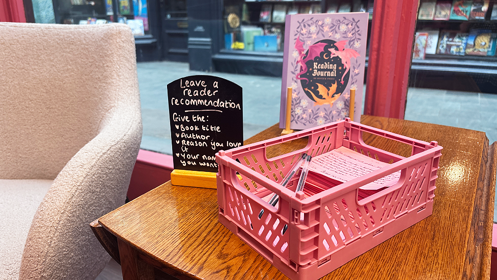 A pink box on top of a side table next to an armchair in the window of Love Stories bookshop. There are cards and pens for people to write recommendations of books. A mini blackboard asking for reader recommendations and instructions - leave the title, author, reason you love it, your name if you want to - is written in chalk.