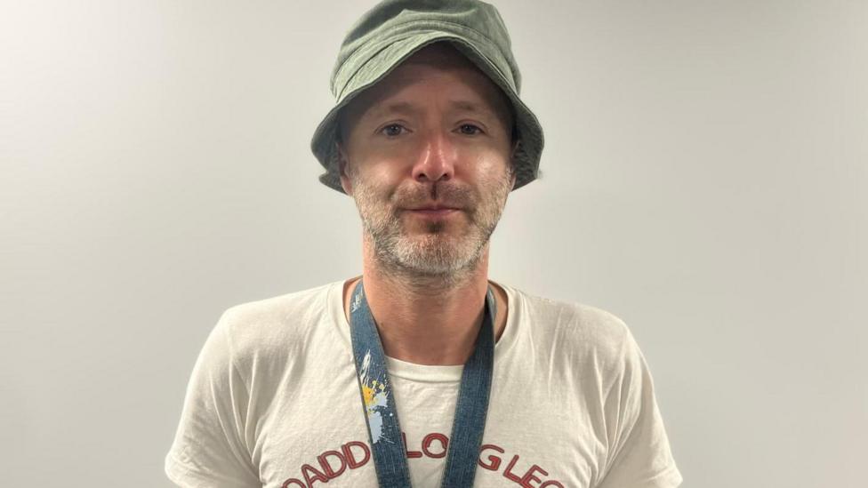 Philip Wallace stands looking directly into the camera. He is wearing a green bucket style hat, white t shirt and a lanyard.