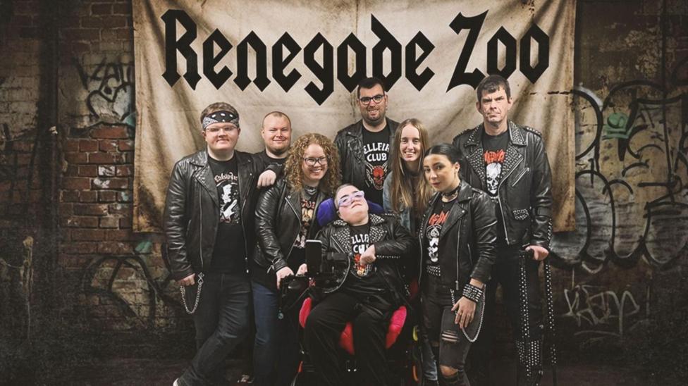 The members of Derry band renegade Zoo are dressed in black leather jackets. There are eight people, four men and four women. In centre of the pic is a woman in a wheelchair. The others are stood around her. In the background is a banner with the band's name written on it