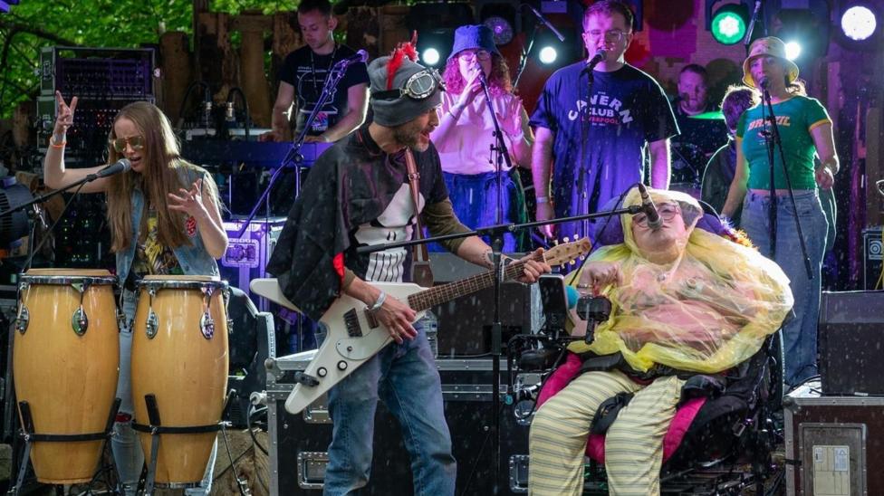 The band are performing on stage at a festival. The young woman in the right of the picture is singing into a microphone. She is wearing a yellow rain poncho and is a wheelchair user. Beside her is a man playing an electric guitar and to his left is another young woman who is playing Conga drums. Behind them other young people are singing into microphones and playing instruments.