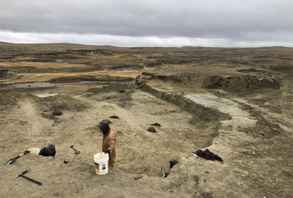 A wide view shows the barren brown landscape of the Badlands in South Dakota, with two people in the foreground on a dig with tools around them, on a cloudy grey day.
