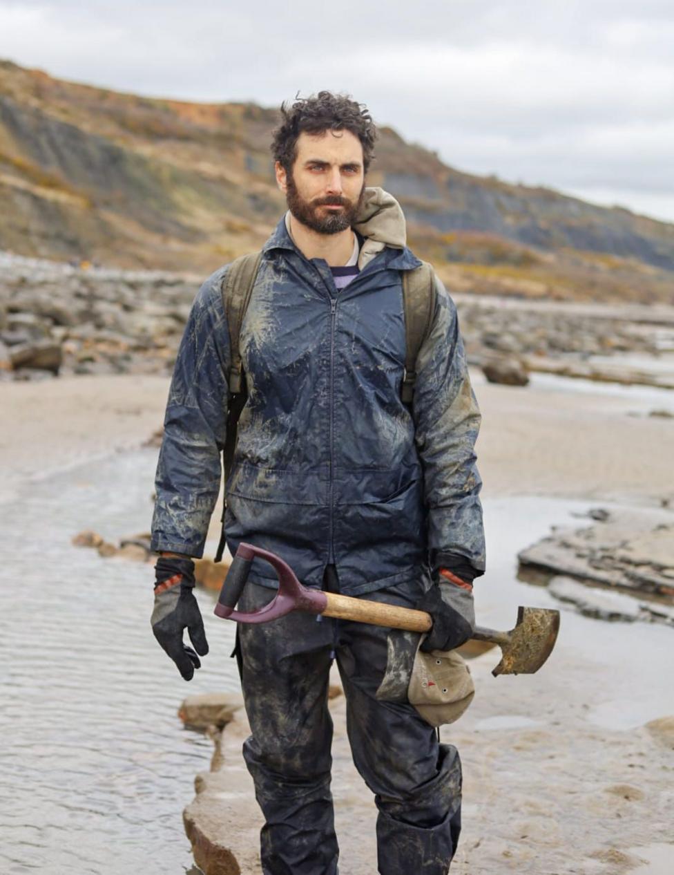 Dr Fiann Smithwick, a man with black hair and a beard, stands on stone next to water with a hillside in the background on a grey cloudy day.