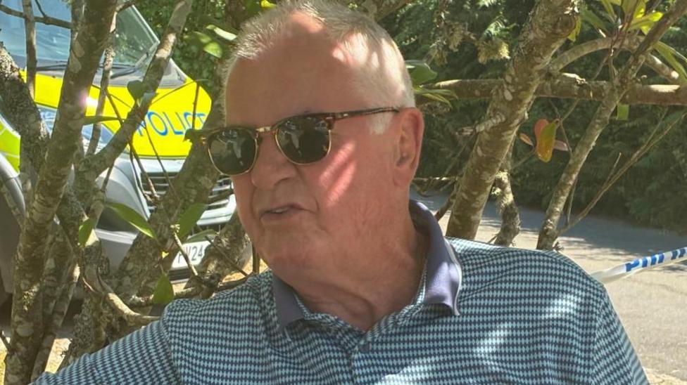 Pete Horrall, who has grey hair, wears a blue patterned polo shirt and sunglasses as he speaks to reporters. A police van and police tape can be seen behind him