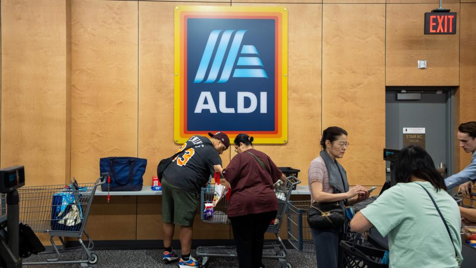 An Aldi logo sign hangs above a man in a blue jersey with yellow numbers, green shorts, and a backwards cap as he removes items from a cart and a woman with a burgundy shirt and black pants stands beside him. A woman in a pink shirt with a grey sweatshirt looks at her phone. Meanwhile, a woman with a green shirt removes items from a grocery cart.