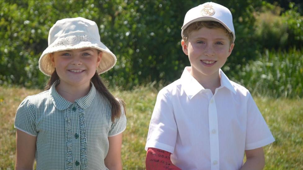 A young girl and boy smile at the camera. They are both wearing school uniform and sun hats and are standing against a backdrop of grass and a riverbed.