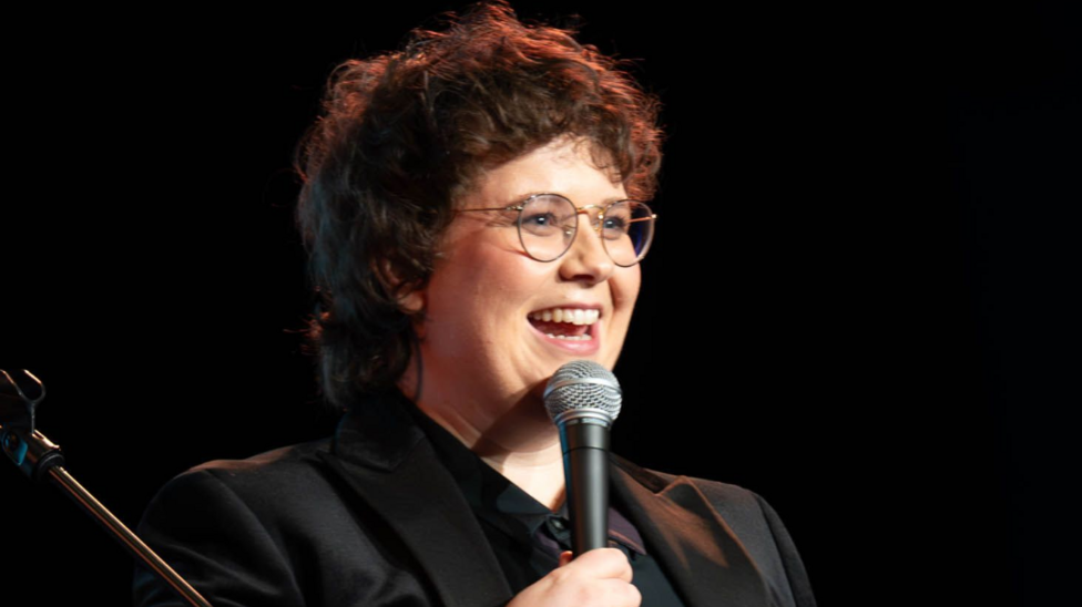 Emer Maguire is performing on stage. She is hllding a miucrophone, is wearing a dark shirt and jacket, and has dark hair. She is wearing glasses.