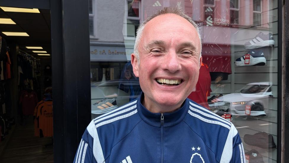 Raymond McGoldrick is wearing a football tracksuit as he stands in front of a shop window. He has short greying hair and is smiling.