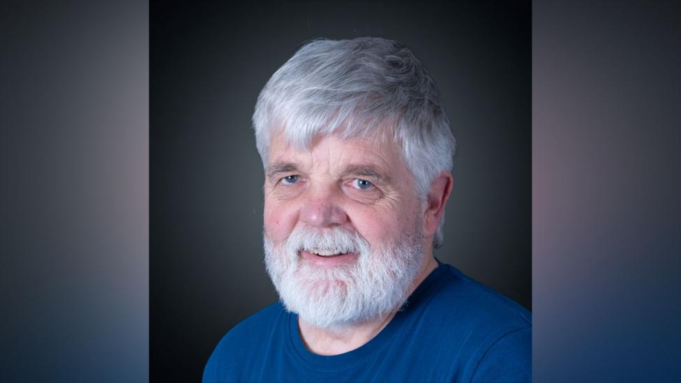 A headshot of an older man with a white beard and white hair. He is wearing a dark blue top.