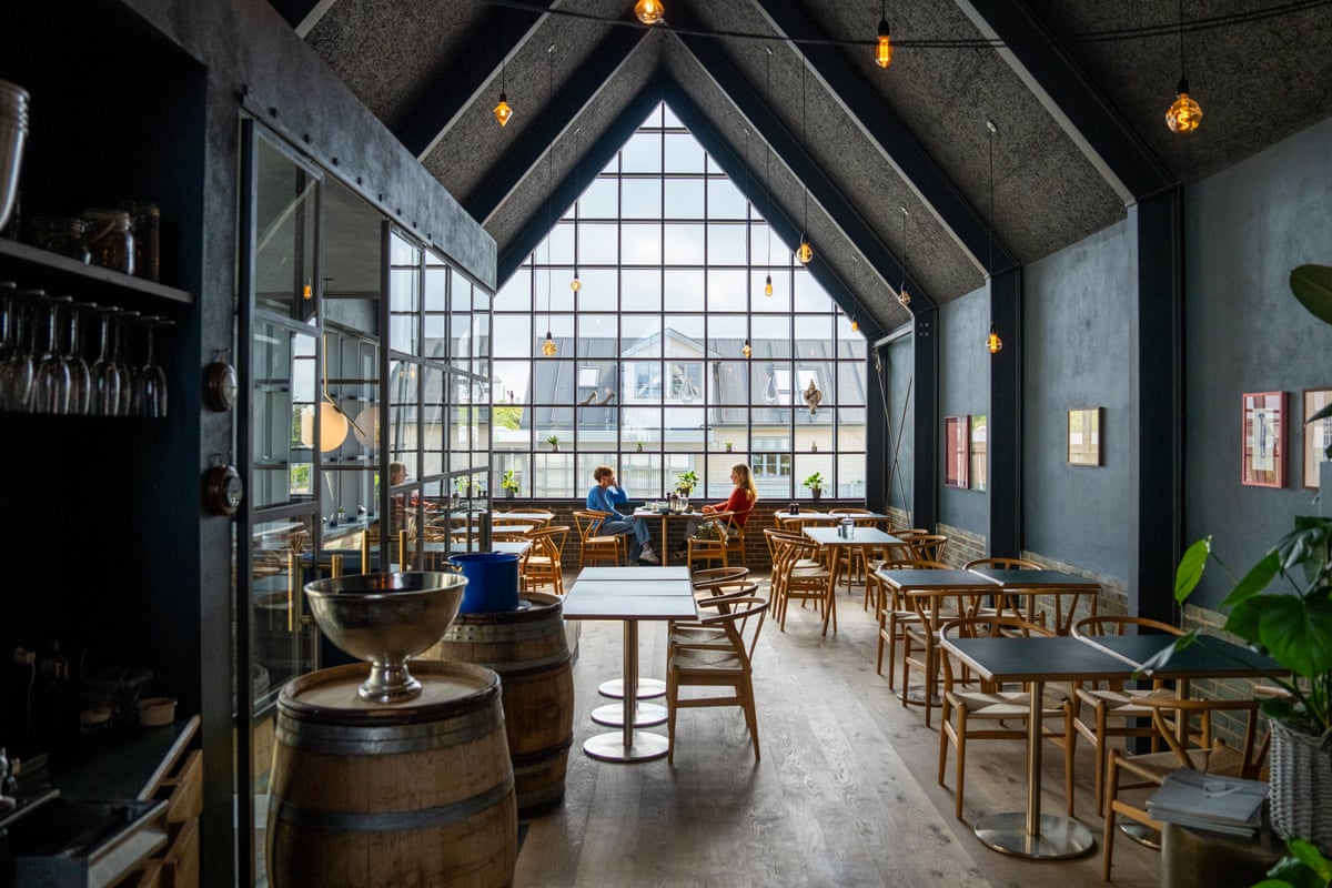 High-ceilinged restaurant with wooden tables and chairs and a front composed entirely of window panes