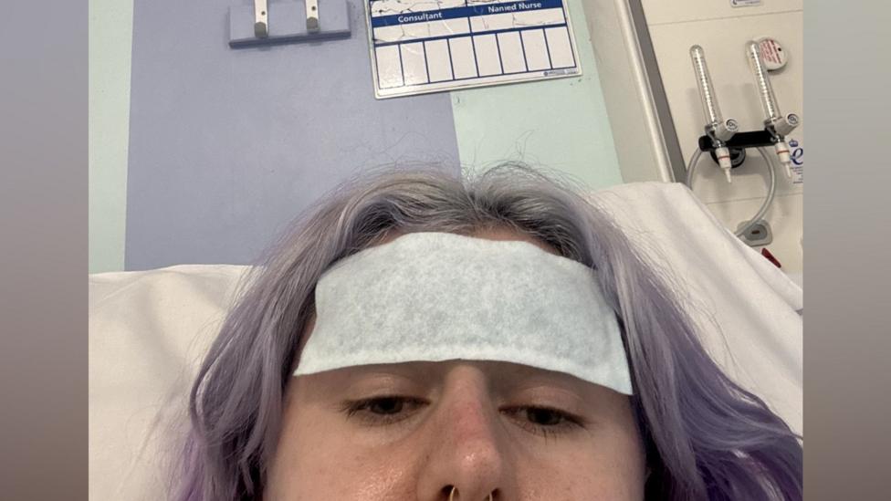 A young woman with short, light purple hair lies in a hospital bed with a patch on her head.