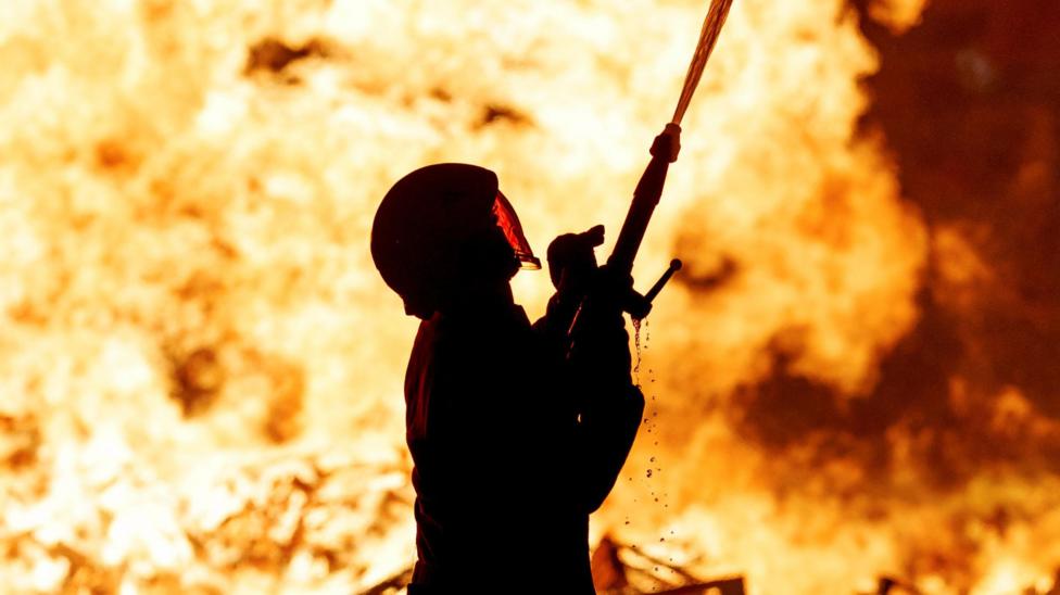 A firefighter in fireproof gear, including an enclosed helmet, sprays a jet of water from his hose into the air. Behind his flames explode.