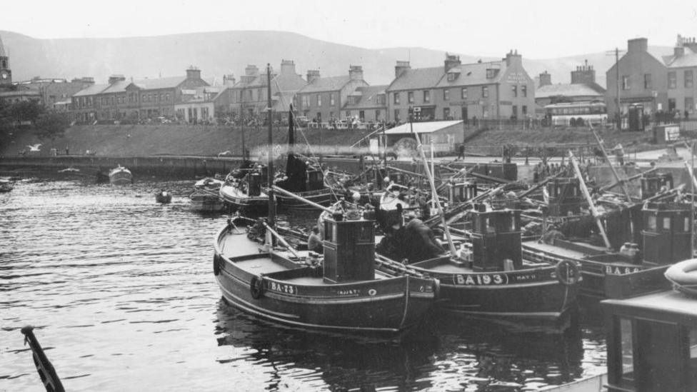 A black and white picture of Girvan, with rows of fishing boats docked by the pier