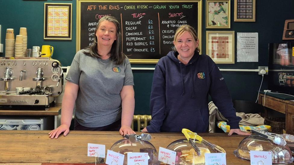 Two women stand behind the counter of a cafe, with an array of cakes for sale. 