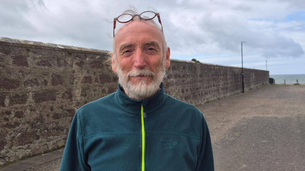 Chris Saunders , a man with a white beard and a aqua top , standing on a street near the seafront in Girvan
