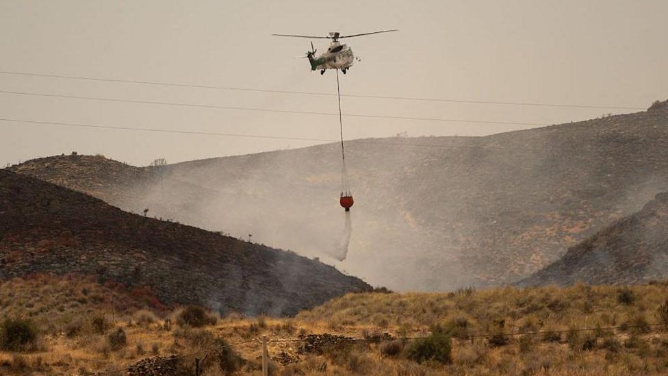 A wide angle photo showing a helicopter dropping water on a grass and shrub wildfire in Spain.