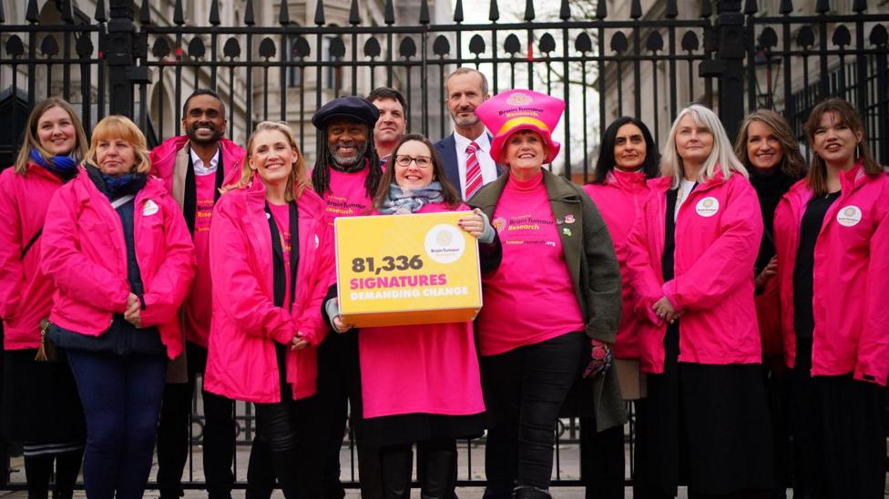 A group of 13 people stand by the gates for Downing Street wearing pink jackets and Theo Burrell is in the middle holding a yellow box that reads