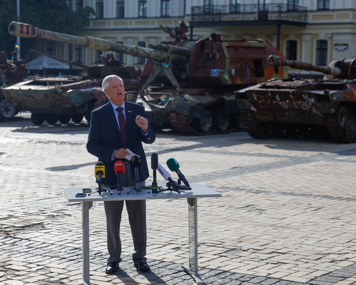 Lindsey Graham speaks at table with microphones on it in a square with tanks in the background