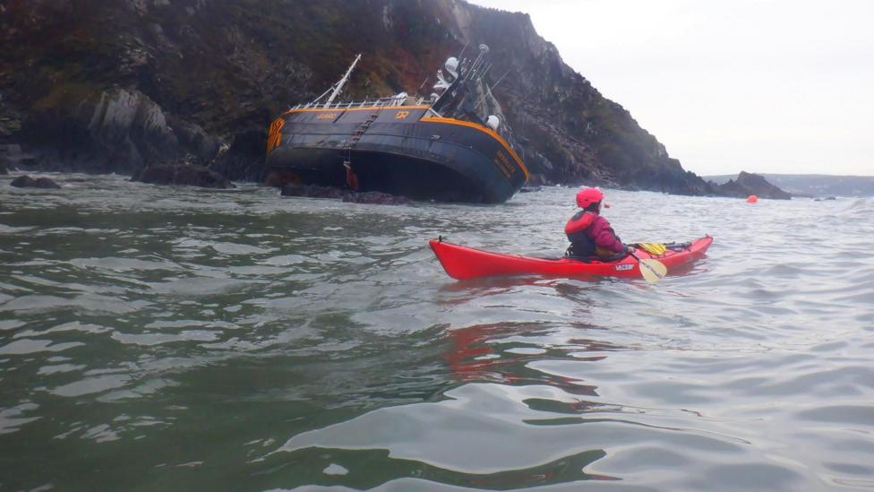 Resolute on the rocks in the background, tilting to one side, dark blue hull with some orange near the top, a kayaker in the foreground