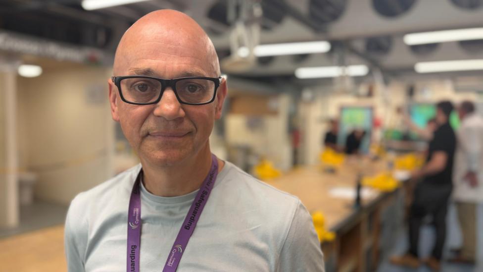 A man wearing a light t-shirt and purple lanyard in a workshop with students working in the distance, behind him.