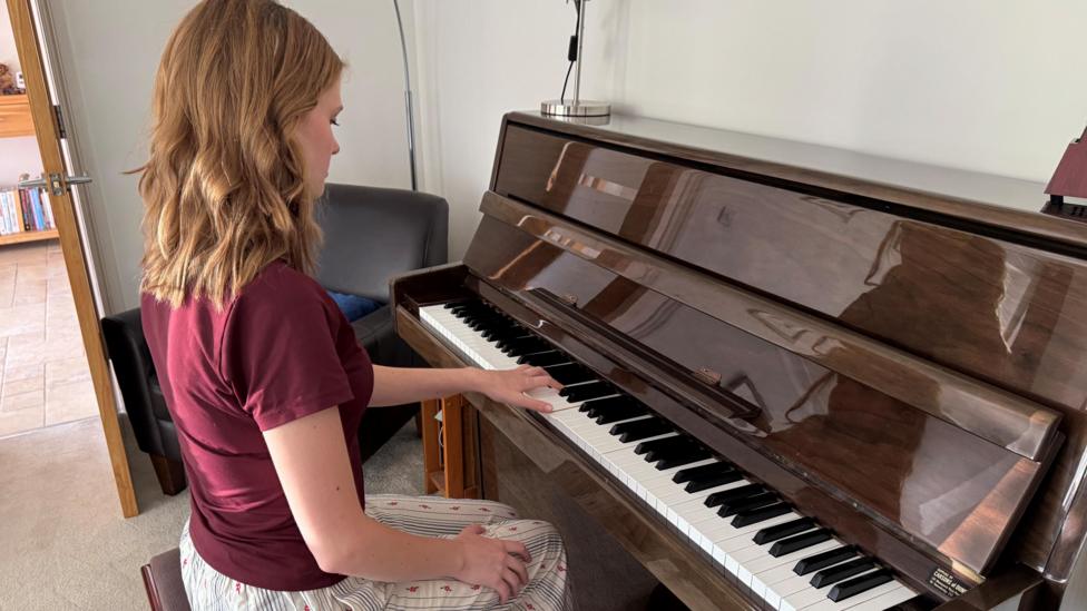 Freya is sitting at a glossy brown piano, with her left hand resting on the keys. Her right hand is resting on her knee, which is where it normally remains while she is performing. She is sitting upright on the piano stool and is wearing a burgundy t-shirt and white trousers with thin blue stripes and a pink dotted pattern. 