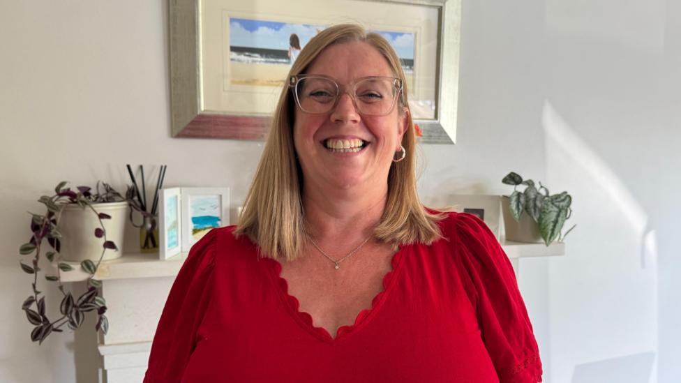Susan Terris has straight, shoulder-length, blonde hair. She is smiling and wearing large clear-framed glasses. One hoop earring can be seen on her right ear as her hair is tucked behind it. She is also wearing a delicate silver necklace and a red top with ruffled edge around the neck. She is standing in front of a white wall which is decorated with a number of paintings and plants sitting on a fireplace mantlepiece.