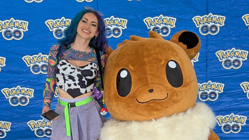 A picture of the Pokémon GO content creator j0beats who has blue hair, and is smiling next to a large costumed Eevee Pokémon against a blue backdrop which says Pokémon Go. She is wearing a black and white cow style top and a denim skirt.