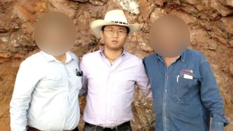 A man wearing a white cowboy-style hat and a pale purple shirt tucked into his trousers. He is wearing glasses and a necklace and is looking at the camera. There is a man on his left and another on his right - the faces of these two men are blurred to obscure their identities. 