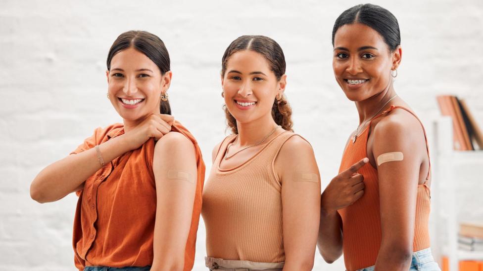 Three smiling young women stand together. Each has just been vaccinated and has a sticking plaster on the injection site. They have rolled up their sleeves to show the camera