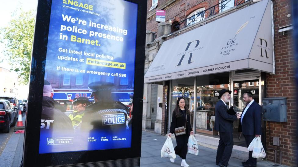 A streetside advertising display promotes increased police presence in Barnet, standing beside a shopfront on a busy urban high street. Several pedestrians carrying shopping bags walk past the storefront, while parked vehicles and brick buildings line the street in the background.