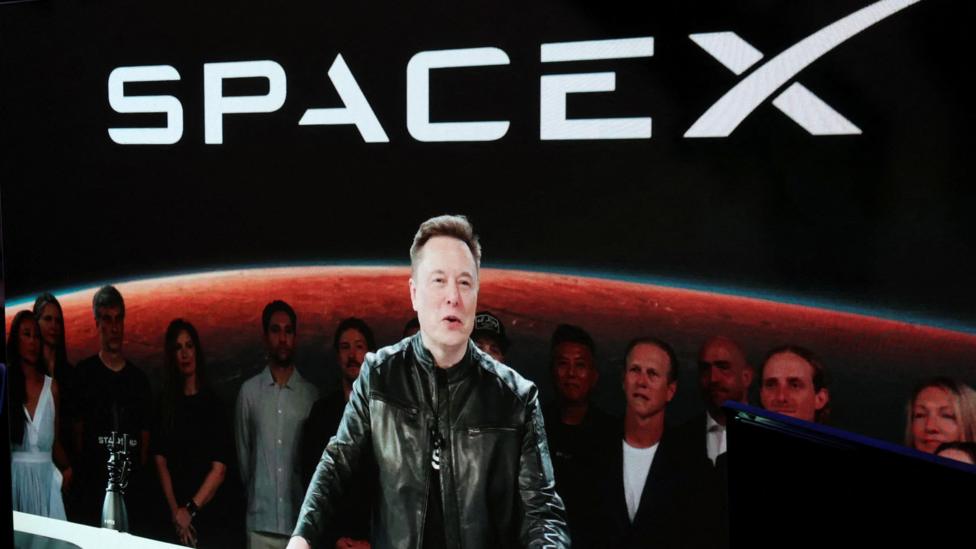 Elon Musk appearing via video stream wearing a black leather jacket an speaking at a podium upon the June debut of his company SpaceX on the Nasdaq stock exchange.
