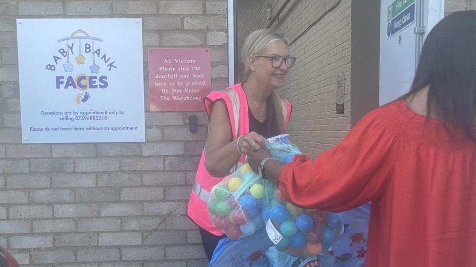 A bag of balls, being given to a woman, in a pink high-vis jacket, by an open door in a brick building. You can see the back of a woman in a red dress.