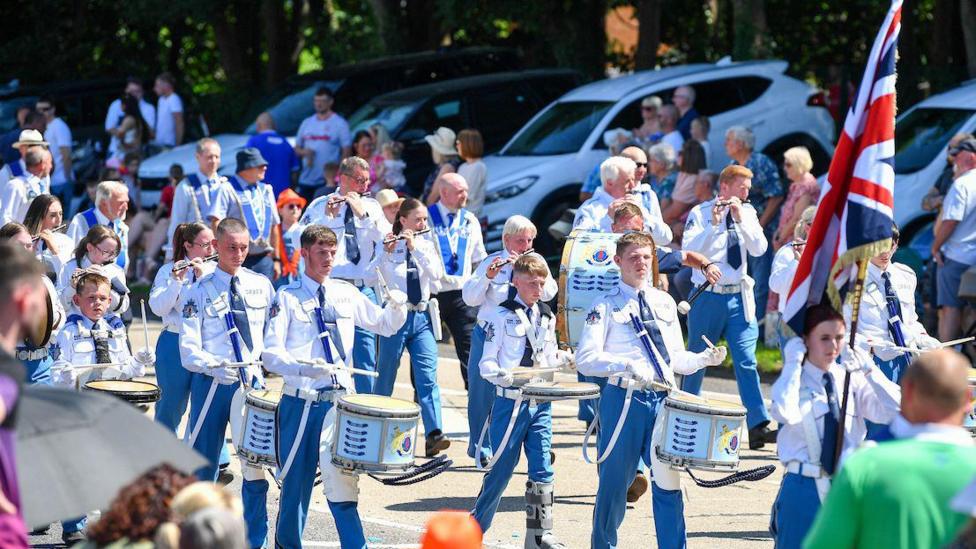 A large band holding drums wearing white shirts and blue trousers.