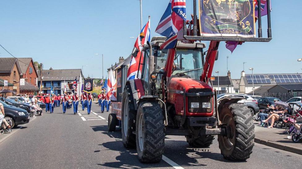A red tractor is at the front of a parade draped in Union flags.