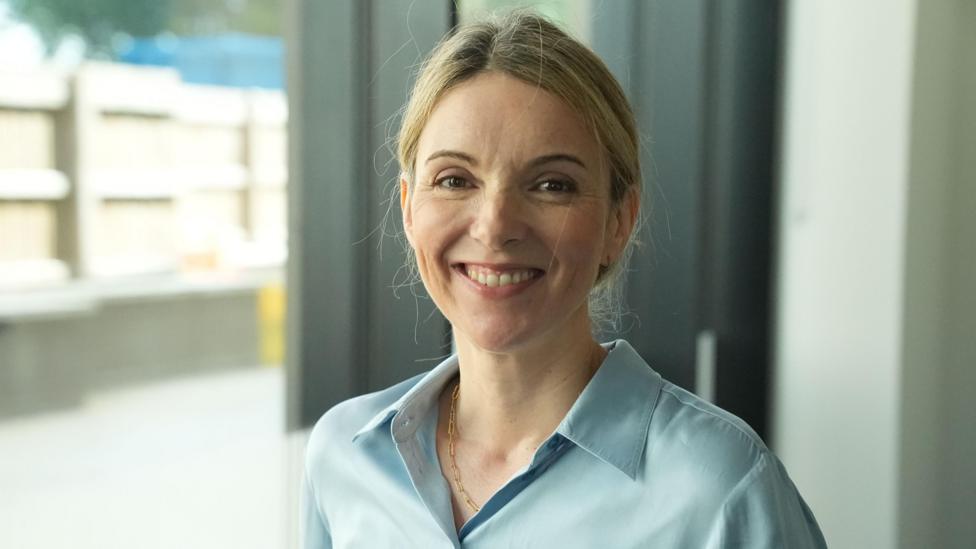 The image shows a person standing indoors near large windows in a bright, modern setting. The person is wearing a light blue button-up shirt and a thin necklace, with hair pulled back away from the face. The photograph is framed as a head-and-shoulders portrait, with the subject positioned prominently in the foreground. The background is softly blurred and includes window frames, a curtain, and an outdoor area visible through the glass. Natural light illuminates the scene, creating a clean and professional appearance with soft shadows and neutral colors.