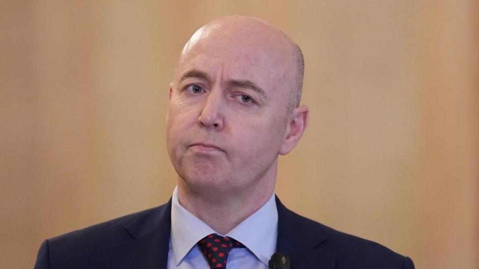 Jon Burrows is bald on top with cropped hair at the side and is wearing a dark suit