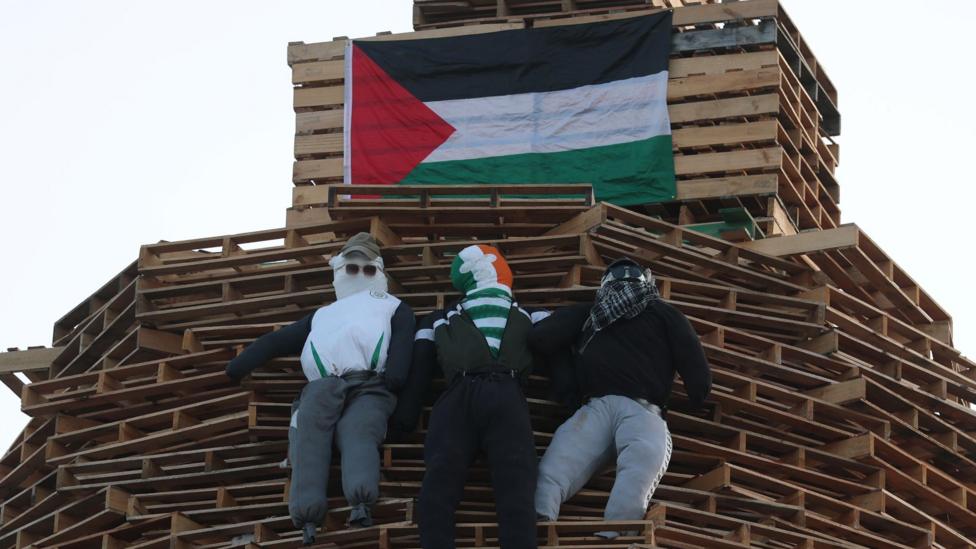 Three stuffed models are pinned to a bonfire, wearing tracksuits and various headwear - including a peaked caps and a green, white and orange balaclava. 