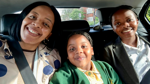 A woman and two young daughters sit together in the back seat of a car. All are smiling.