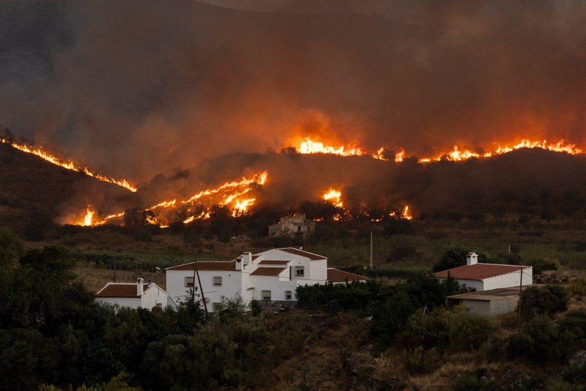 Smoke blots out the sky as flames run the full length of a hilly landscape, with white-walled Mediterranean homes dangerously close by