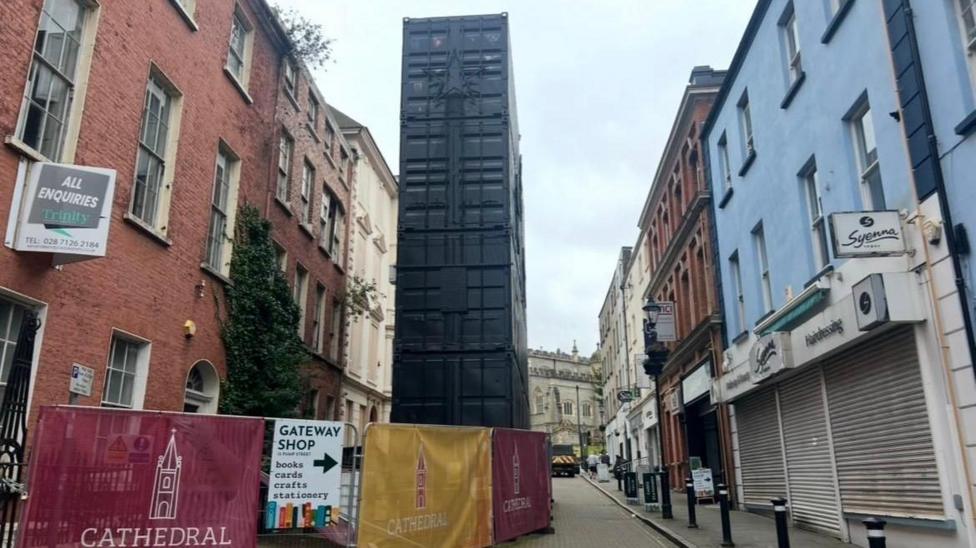 large shipping containers stand upright in a narrow pedestrianised street. Shops line both sides of the street. A number of crash barriers are in front of the container.